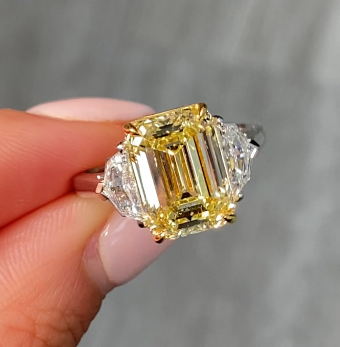 A natural yellow emerald-cut diamond ring held between fingers against a soft gray blurred background.
