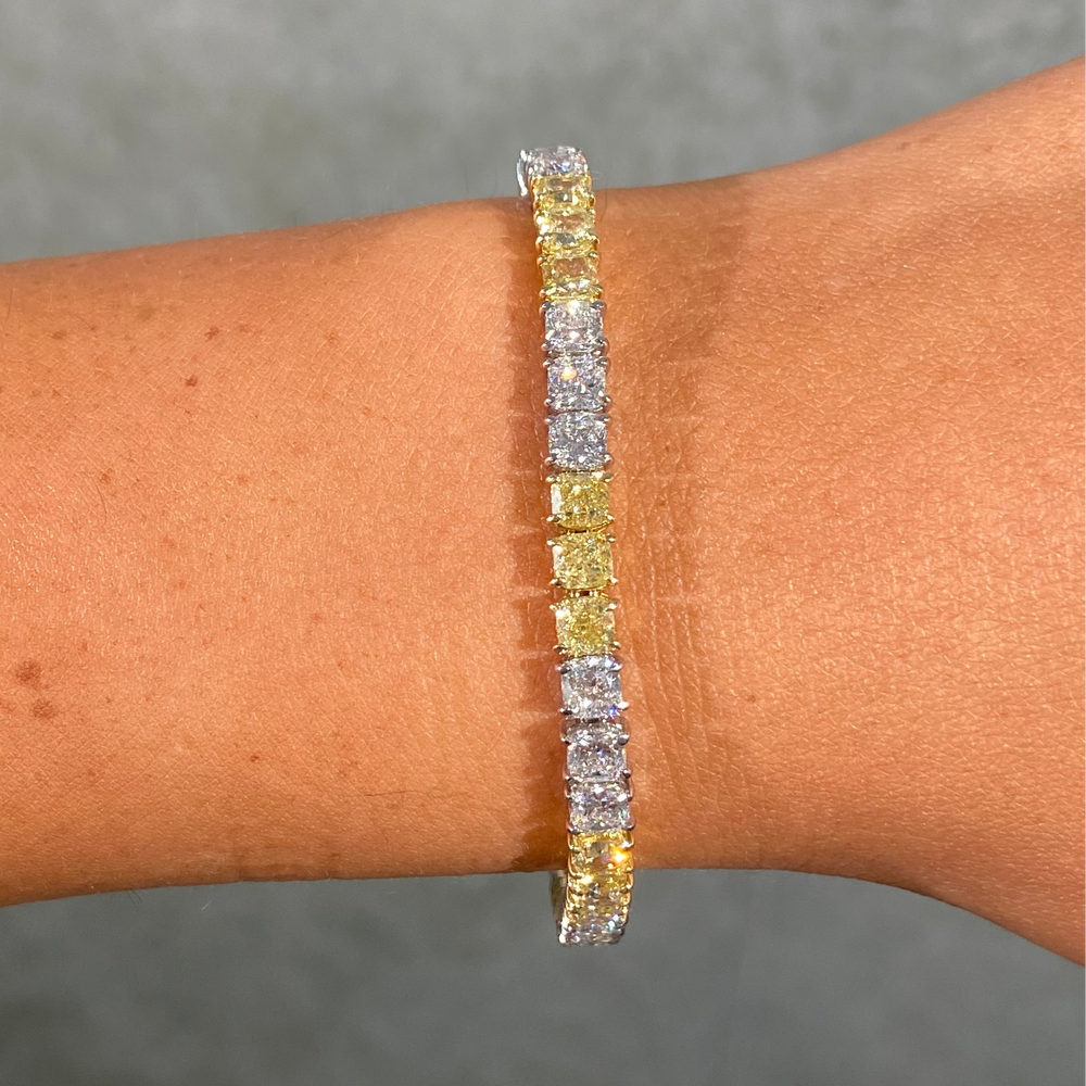 A close-up of a tennis bracelet worn on a wrist. The bracelet features alternating yellow and clear stones set in yellow gold, photographed against a textured gray background.